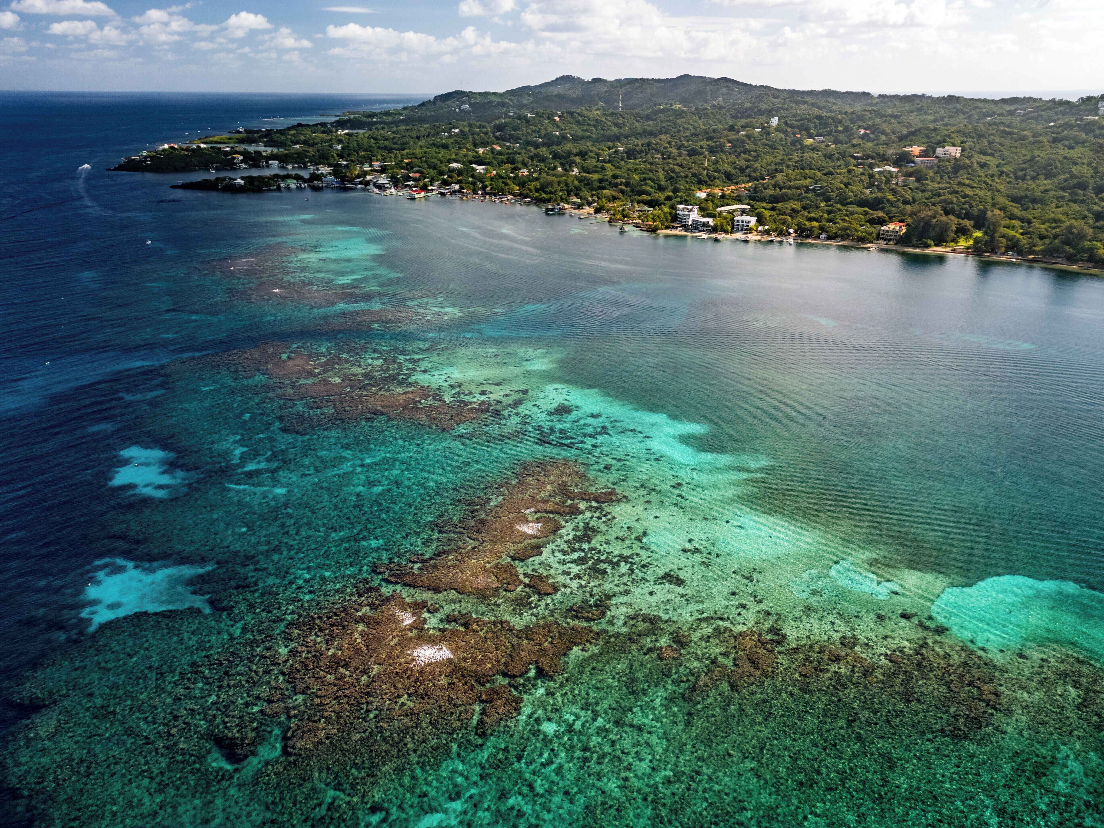An aerial view of a coastal area with clear turquoise waters, a small island with buildings, and a mountainous backdrop.