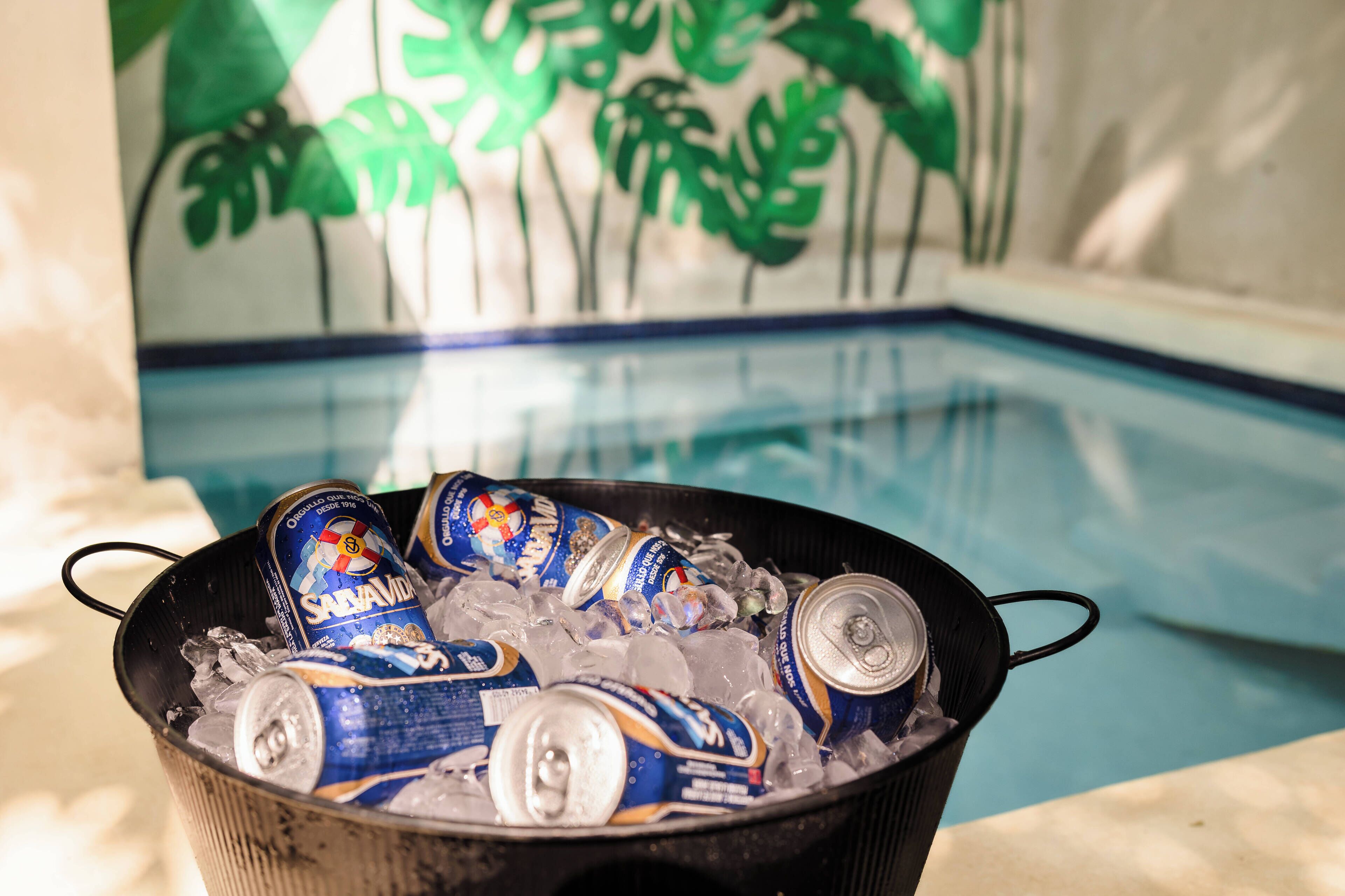 A bucket filled with ice and cans of beer, placed near a pool with a tropical mural in the background.