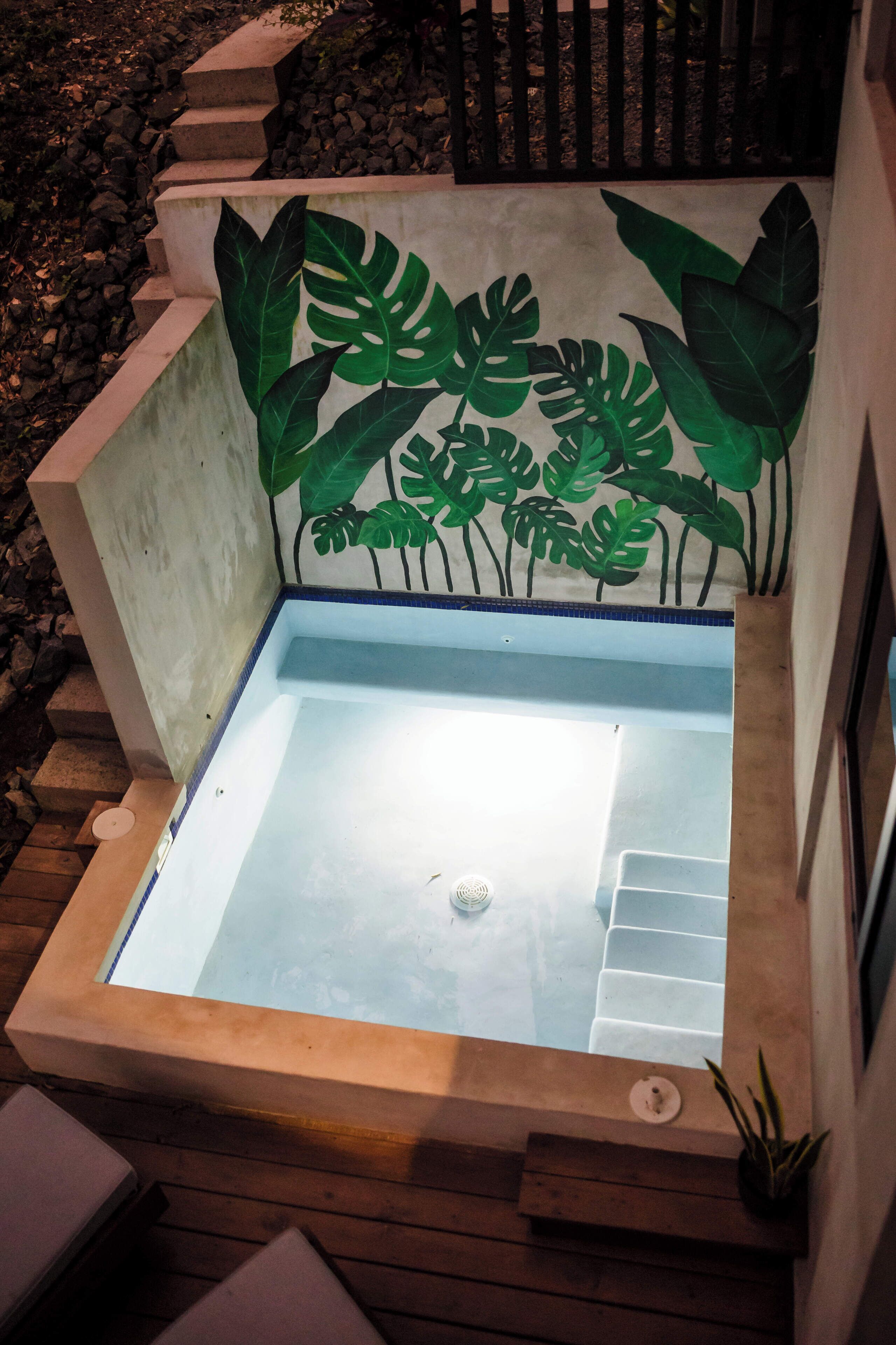 Un jacuzzi con borde de hormigón y un diseño de hoja tropical pintada en la pared.
