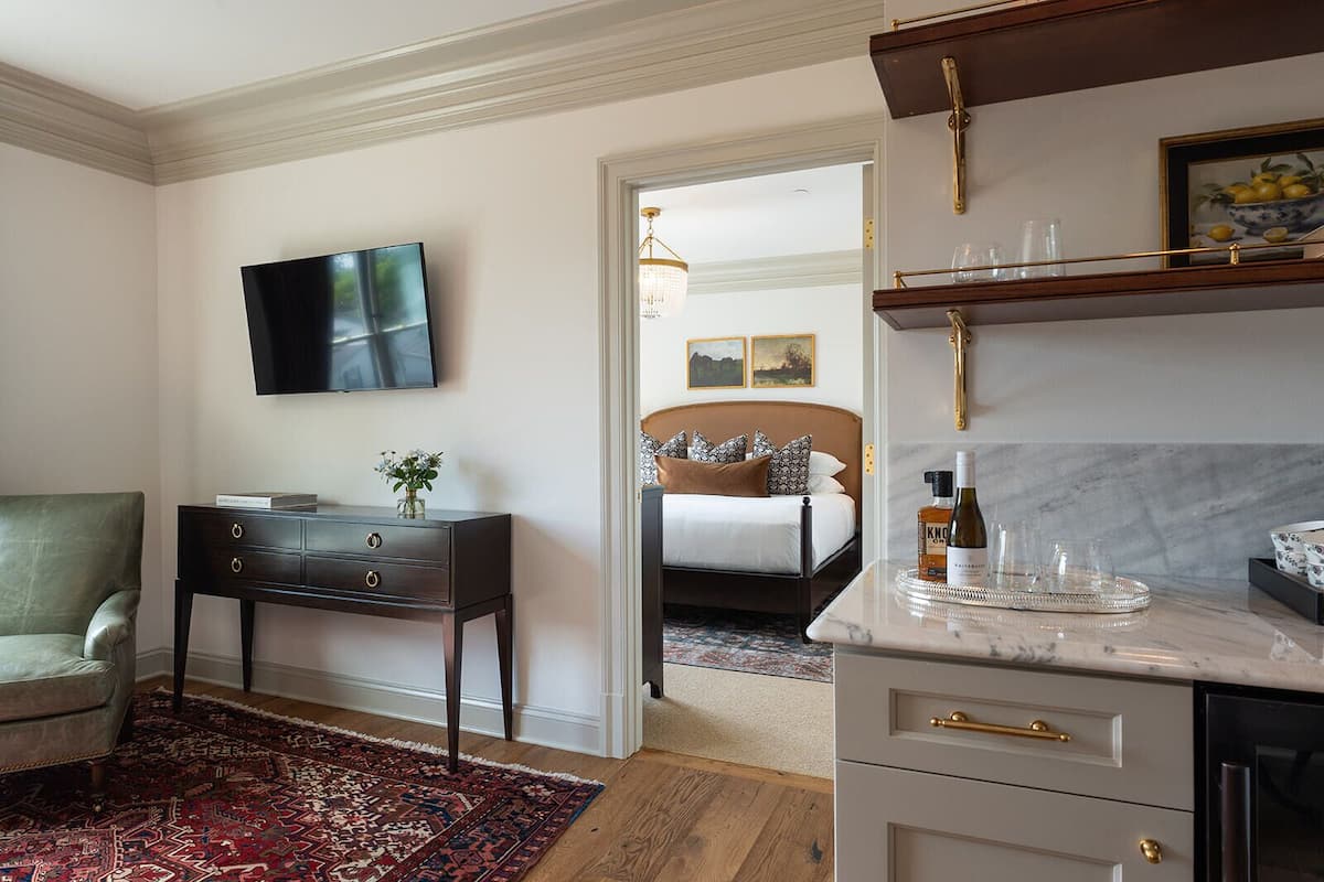 Ett hotellrum med en säng, ett skrivbord, en TV och ett köksutrymme med en bänkskiva i marmor och hyllor.