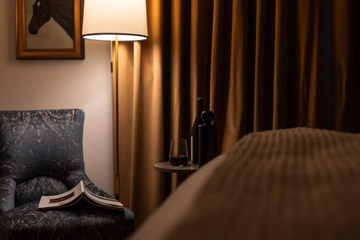 Ett hotellrum med en säng, en stol med en öppen bok, en lampa och ett litet bord med ett glas och en flaska.