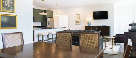 A dining area with a wooden table and chairs, a kitchen in the background, and a flat-screen TV mounted on the wall.