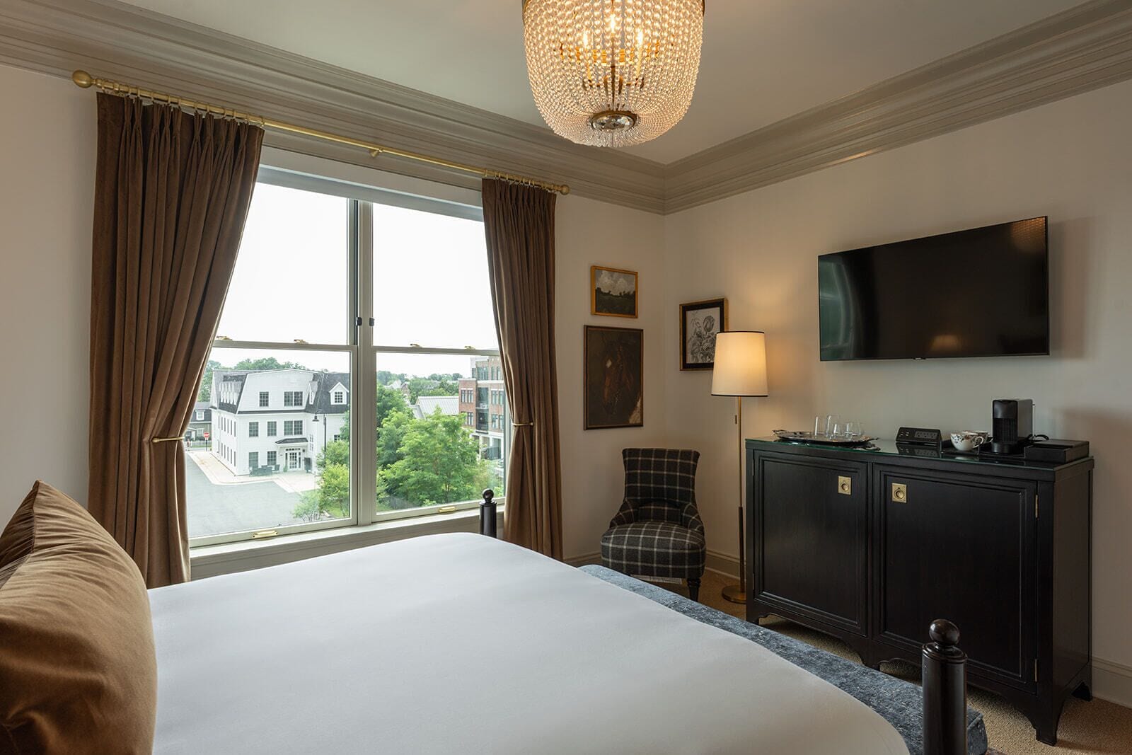 A hotel room with a large bed, a flat-screen TV, a desk with a lamp, and a view of buildings through the window.