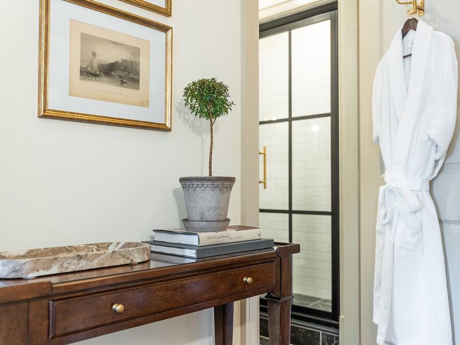 Una consola de madera con una planta en maceta, un cuadro enmarco y una pila de libros. Un albornoz blanco cuelga en una puerta a la derecha.
