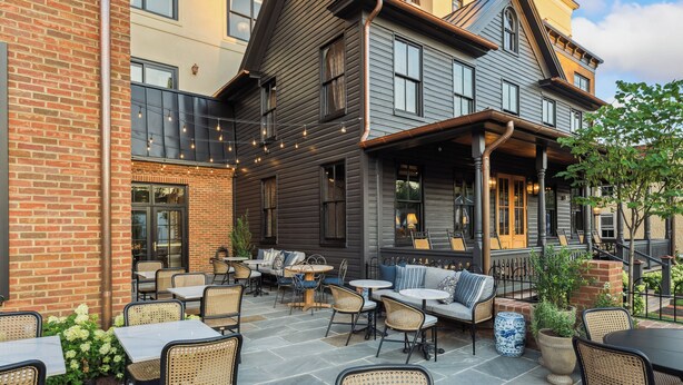 Patio esterno con tavoli e sedie, un muro in mattoni e un edificio con diverse finestre e un portico.