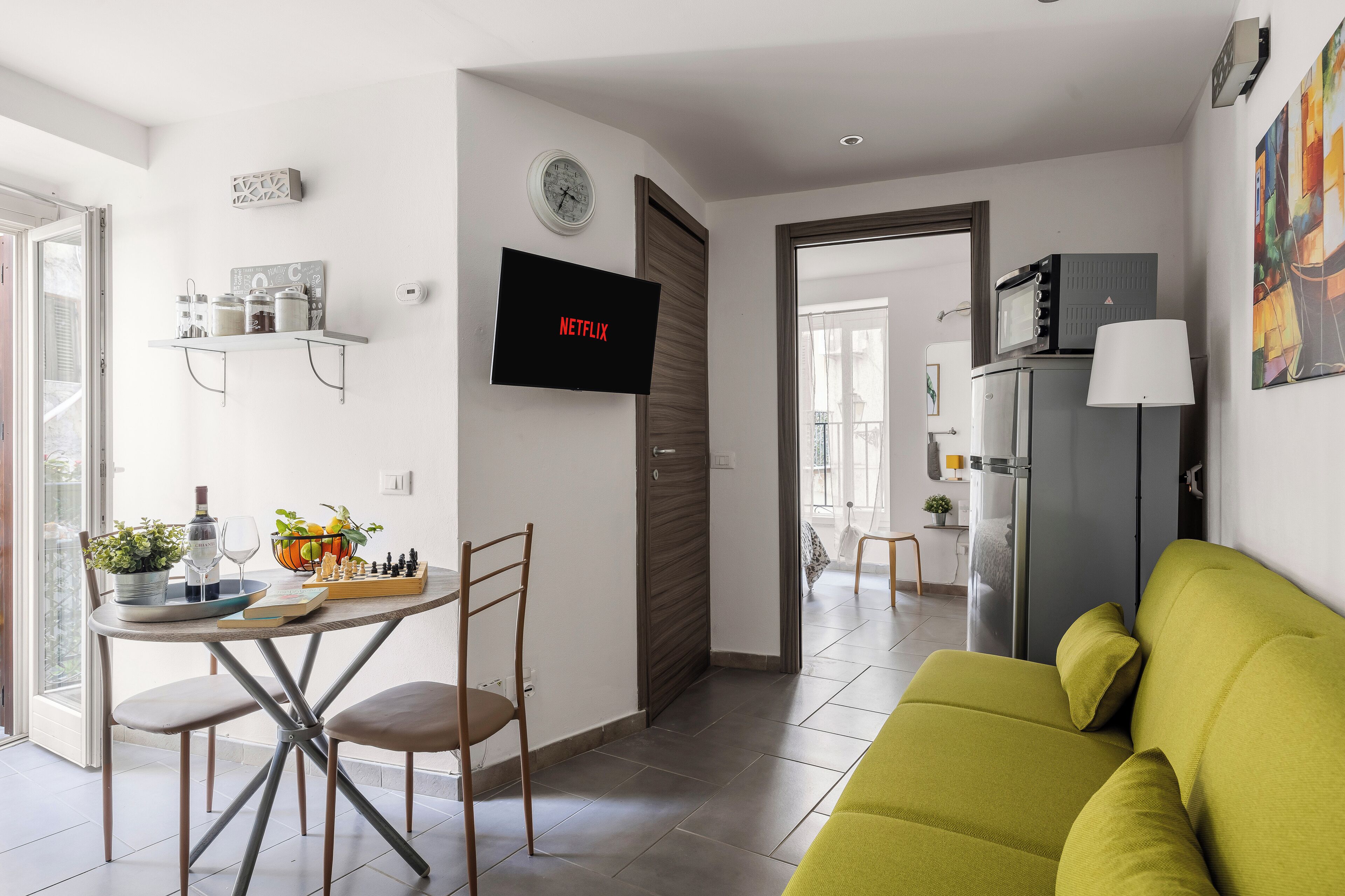 Un salon moderne avec un canapé vert, une table à manger avec des chaises, un téléviseur fixé au mur et un coin cuisine visible à travers une porte ouverte.