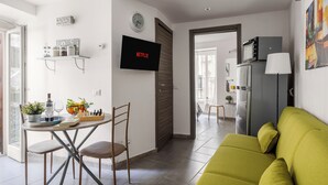 A modern living room with a green sofa, a dining table with chairs, a TV mounted on the wall, and a kitchen area visible through an open door.