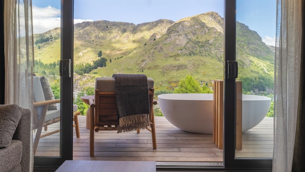 A room with a large window, a wooden deck, a white bathtub, and a chair. - Coronet Ridge Resort (Arthur's Point)