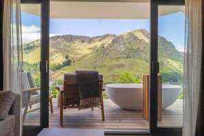 A room with a large window, a wooden deck, a white bathtub, and a chair.