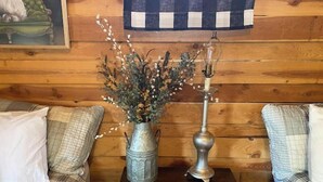 Une pièce chaleureuse avec un mur en bois, un vase en métal contenant des fleurs séchées, une lampe ancienne et un coussin à carreaux posé sur une table de chevet en bois.