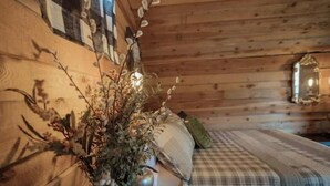 Une pièce chaleureuse avec un plafond en bois, un lit recouvert d’une couette à carreaux, un vase contenant des fleurs séchées et un miroir accroché au mur.
