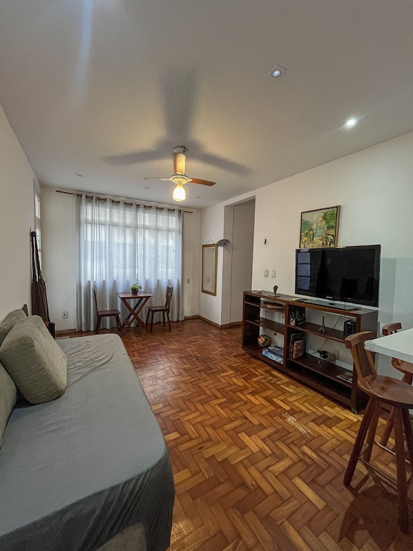 A living room with a sofa, a television, a ceiling fan, and a herringbone-patterned wooden floor.