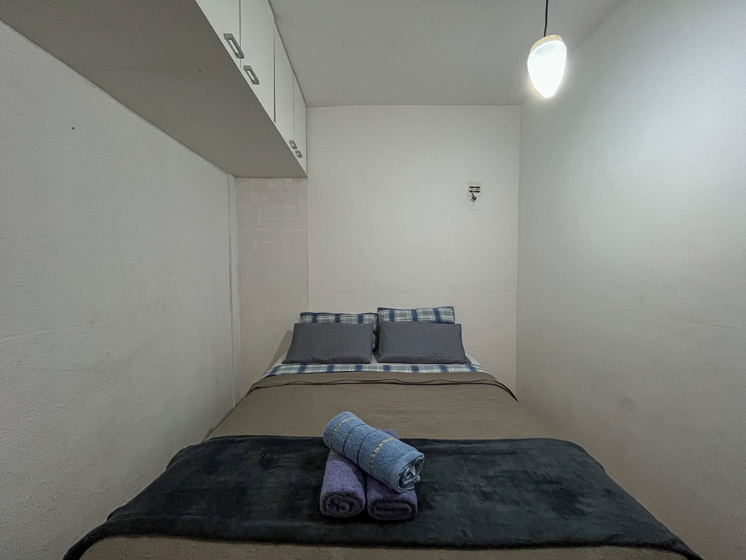 Ein kleiner, schmaler Raum mit einem Bett, einer Wandleuchte und einer Steckdose.