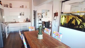 Une cuisine moderne avec des meubles blancs, une table à manger en bois entourée de chaises colorées et un grand aquarium mural.