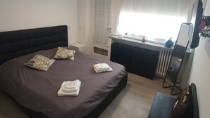 Une chambre à coucher avec un grand lit, une table de chevet, un bureau, un radiateur et une fenêtre avec des rideaux.
