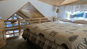 Ein Schlafzimmer mit schrägen Wänden, einem Bett mit gemustertem Überwurf, einem Nachttisch mit Lampe und einem Fenster mit Vorhängen.