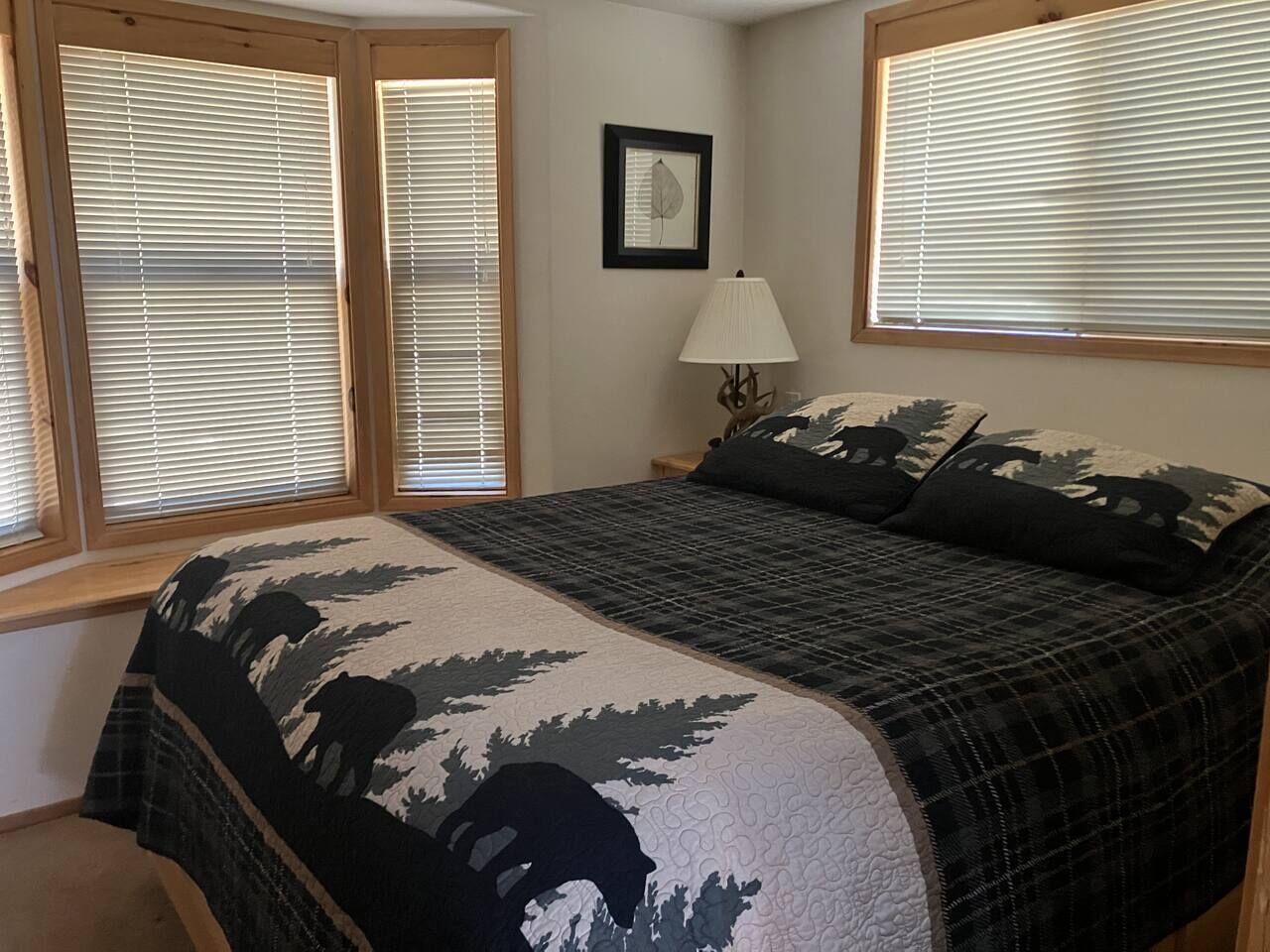 Un dormitorio con cama, mesita de noche, lámpara y dos ventanas con persianas.