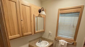 Ein kleines Badezimmer mit Holzschränken, einem weißen Waschbecken, einer Toilette und einem Fenster mit Jalousien.
