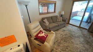 Una sala de estar con sofá, sillón reclinable, lámpara y puerta corrediza de vidrio que da a un espacio exterior.