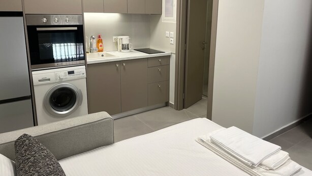 A compact hotel room with a kitchenette, a washing machine, a bed with a pillow, and a neatly made bed with towels. - Petite Fleur Apartment (Lefkada)