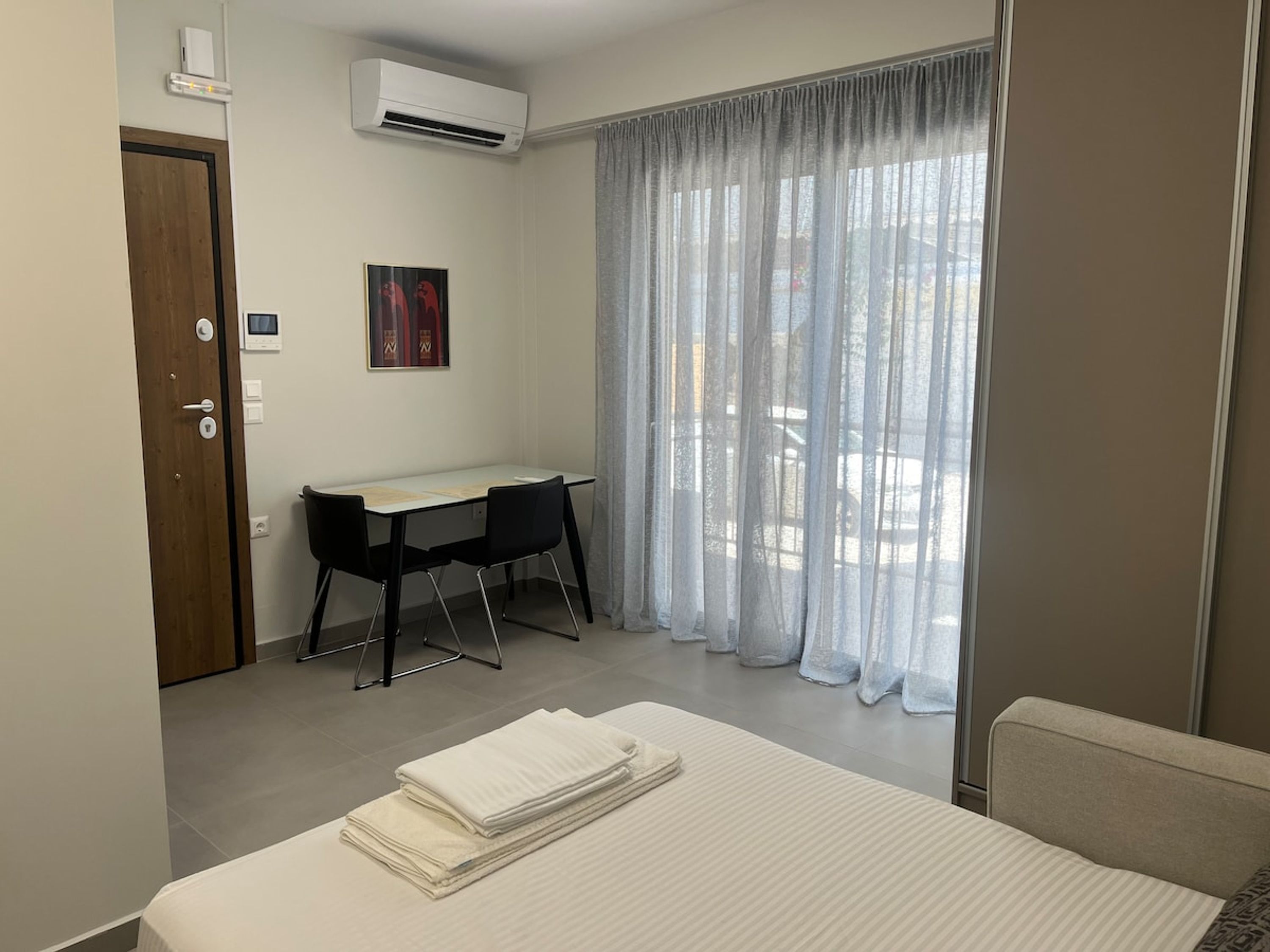A hotel room with a bed, a desk, and a chair. There is a window with curtains, an air conditioning unit, and a door.