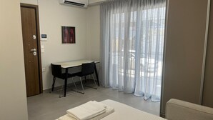 A hotel room with a bed, a desk, and a chair. There is a window with curtains, an air conditioning unit, and a door. - Petite Fleur Apartment (Lefkada)