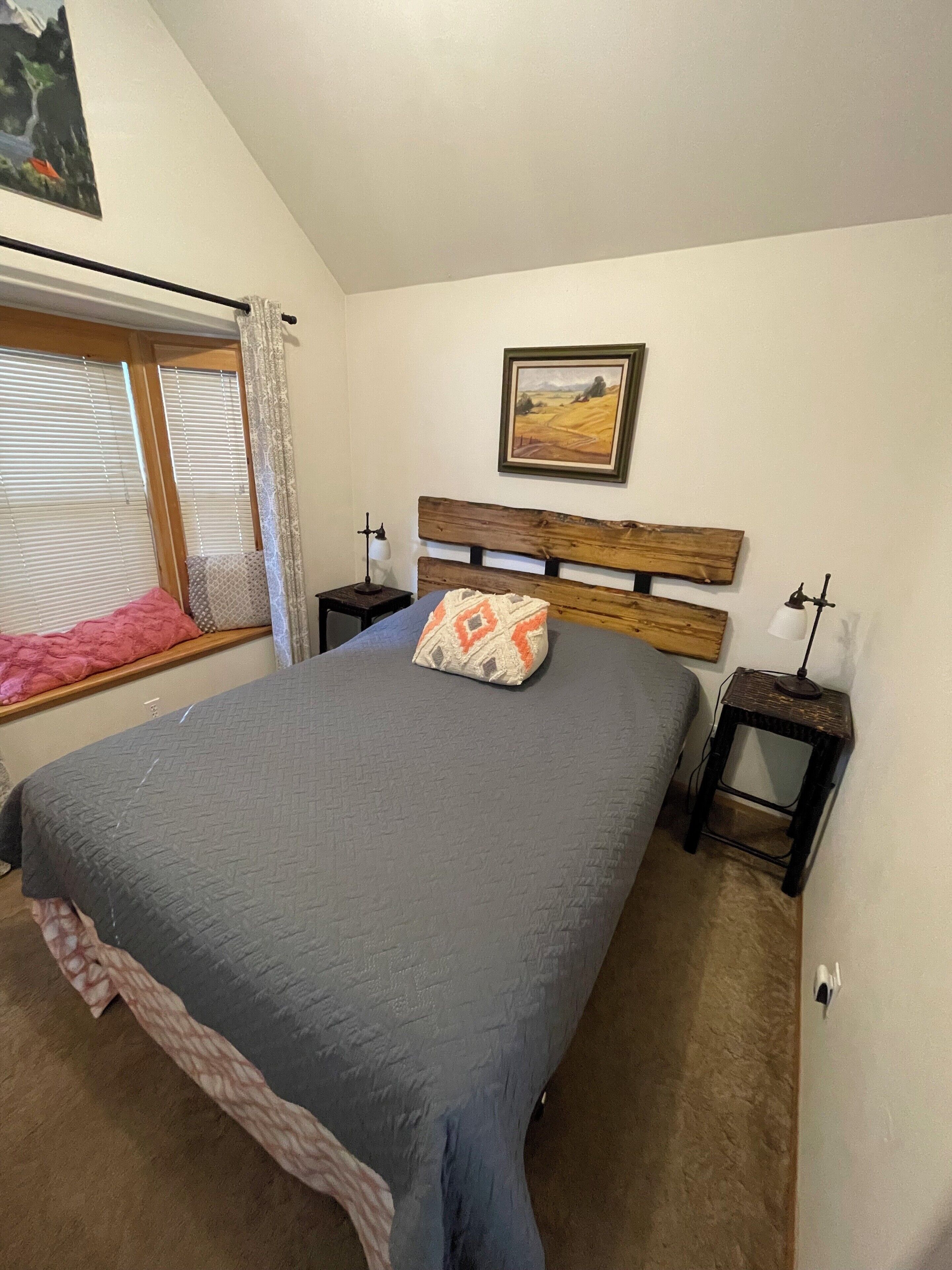 Een slaapkamer met een groot bed, een houten hoofdeinde, een raam met jaloezieën, een nachtkastje met een lamp en een lijst met een afbeelding aan de muur.