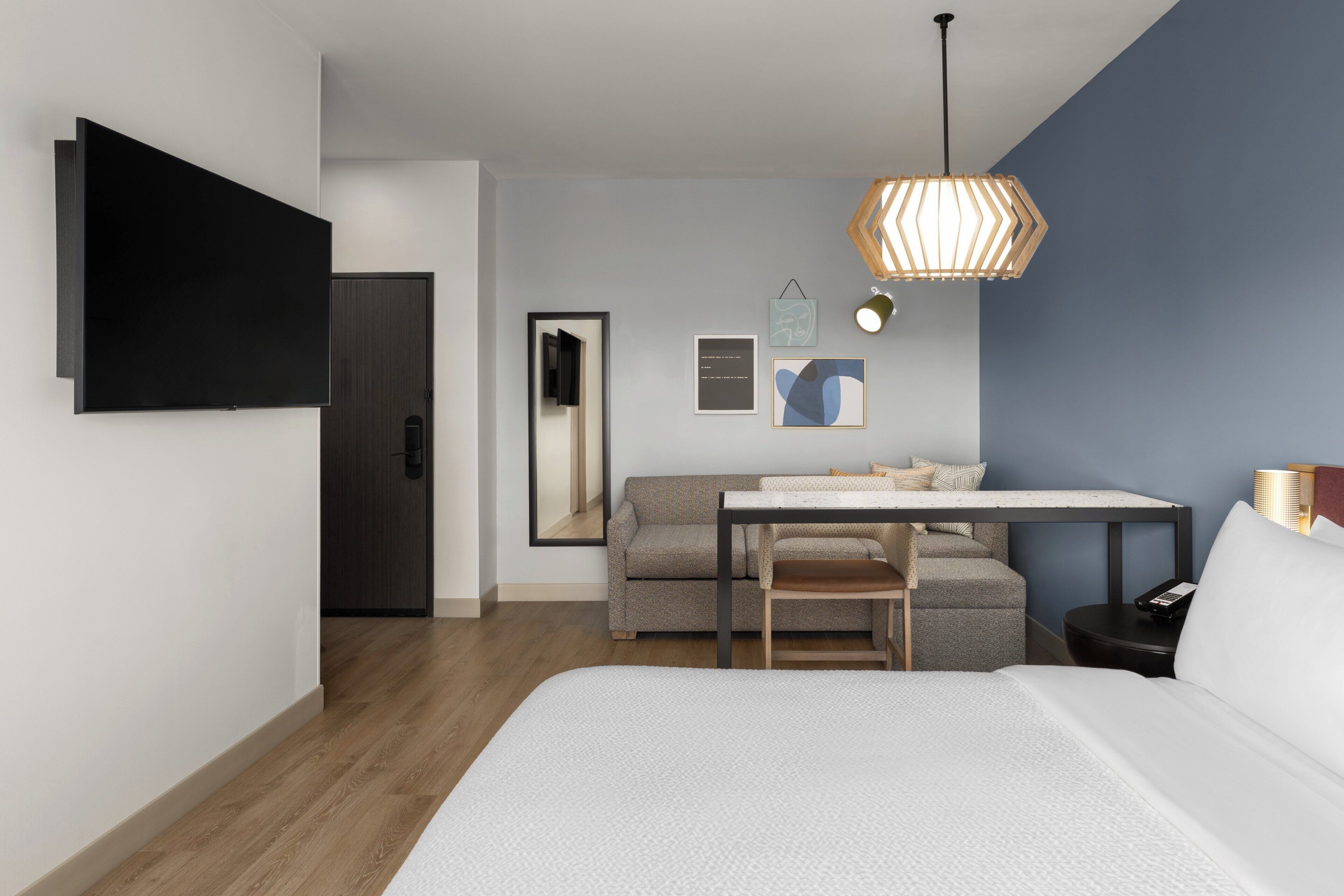 A modern hotel room with a bed, a desk with a chair, a sofa, a TV, and a hanging light fixture.