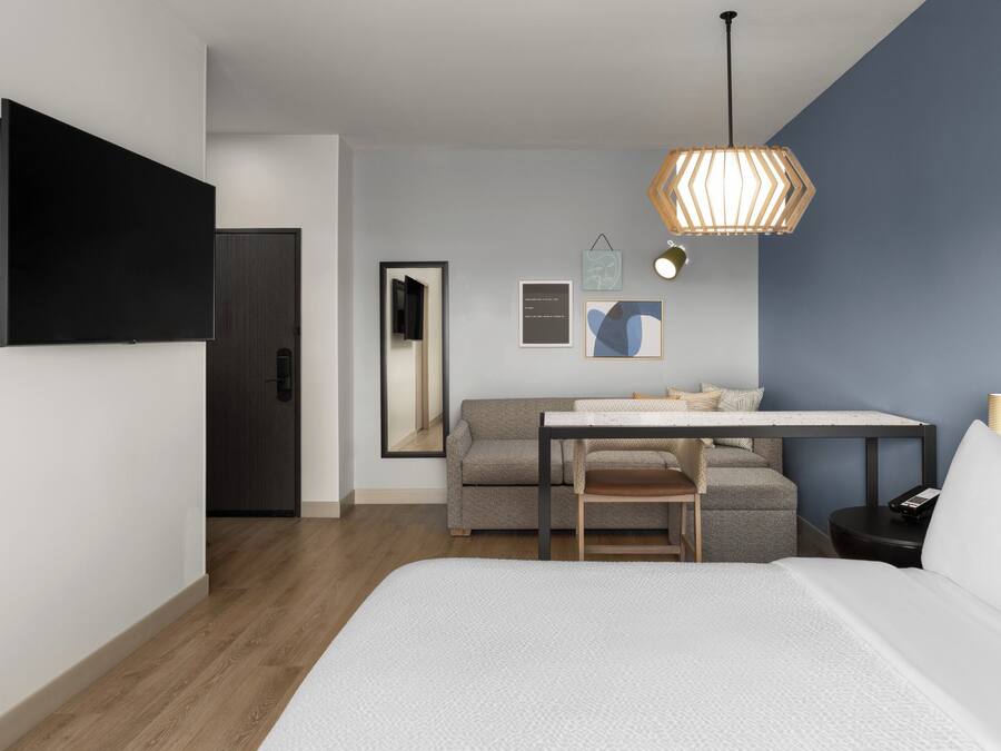 A modern hotel room with a bed, a desk with a chair, a sofa, a TV, and a hanging light fixture.