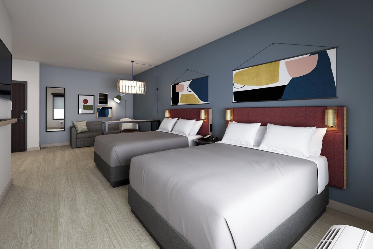 A hotel room with two beds, a desk, and a chair. There are abstract paintings on the walls.