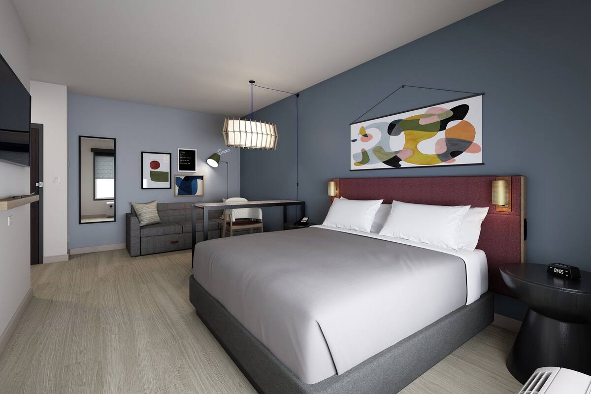 A modern hotel room with a large bed, a desk, and a chair. There is a painting on the wall and a clock on the bedside table.