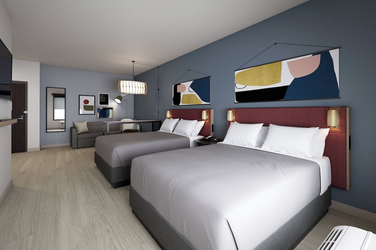 A hotel room with two beds, a desk, and a chair. There are abstract paintings on the walls.