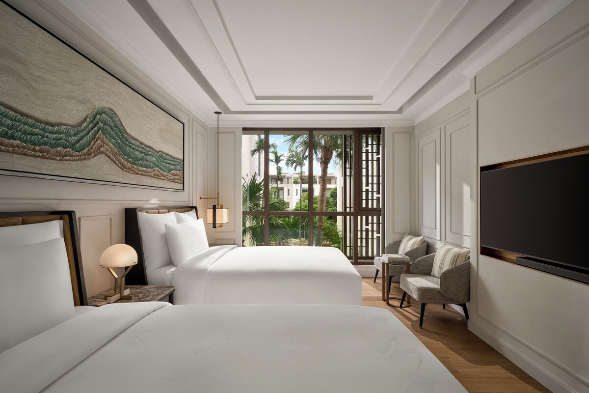 Une chambre d’hôtel dotée d’un grand lit, d’un bureau avec une lampe, d’un coin salon avec des fauteuils et d’une grande fenêtre offrant une vue sur les palmiers et les immeubles.