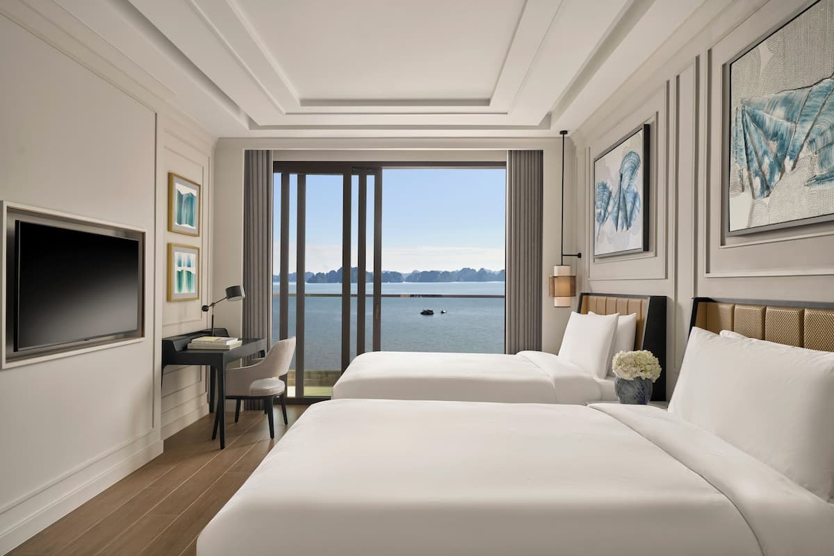Habitación de hotel con dos camas, un escritorio, televisor y ventana grande con vista al agua.