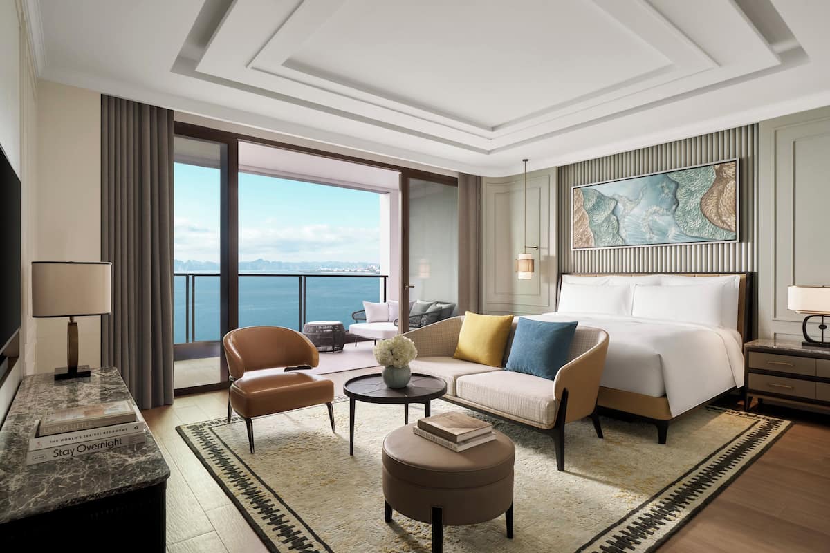 Habitación de hotel con una cama grande, un sofá, dos sillones, una mesa de centro y vista al mar.