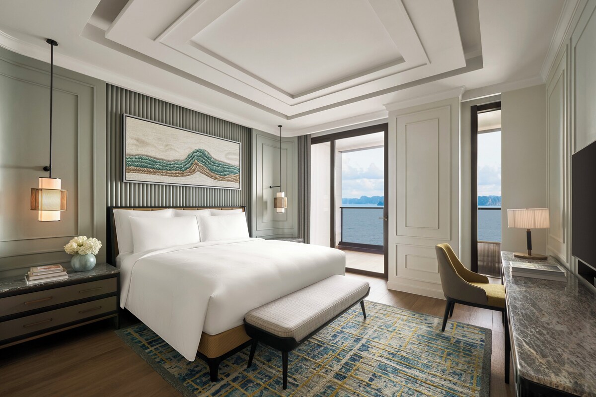 Une chambre d’hôtel avec un grand lit, un bureau, une chaise, une table de chevet, une lampe, un tableau et une vue sur la mer par une grande fenêtre.