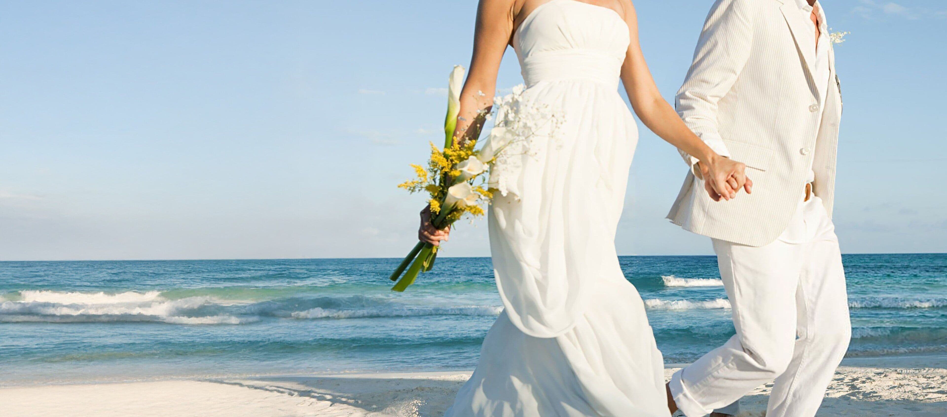 Una pareja camina de la mano por una playa sosteniendo un ramo de flores.