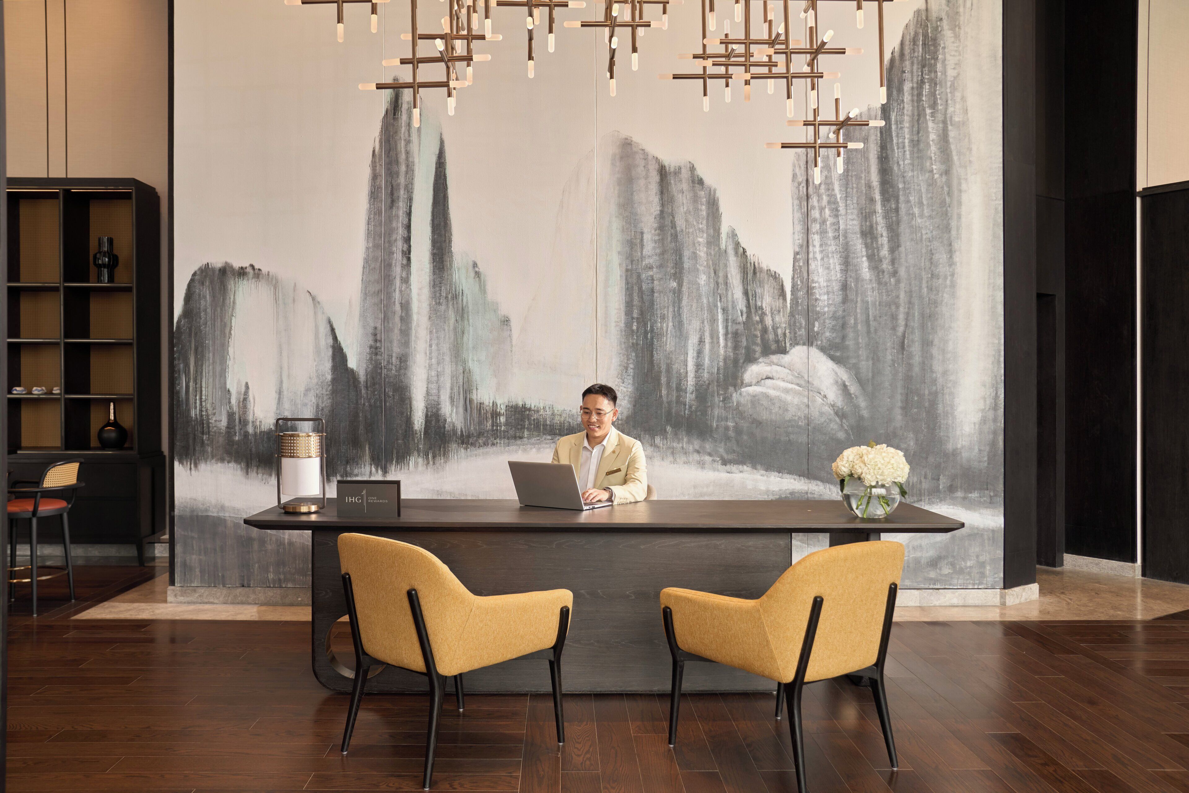 A reception desk with a laptop, two yellow chairs, a vase with flowers, and a large abstract painting in the background.