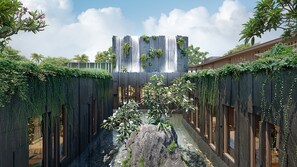 Un étroit sentier bordé de murs en bois et de lianes retombantes, qui mène à un bâtiment agrémenté d’une cascade et de verdure.