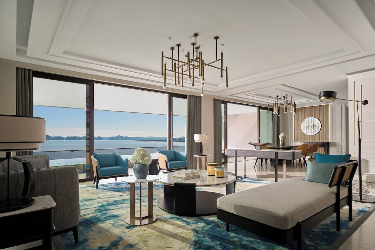Habitación de hotel moderna con un amplio ventanal que ofrece vistas al mar, una zona de estar cómoda con sofá y sillones, una mesa de comedor y una pequeña cocina.