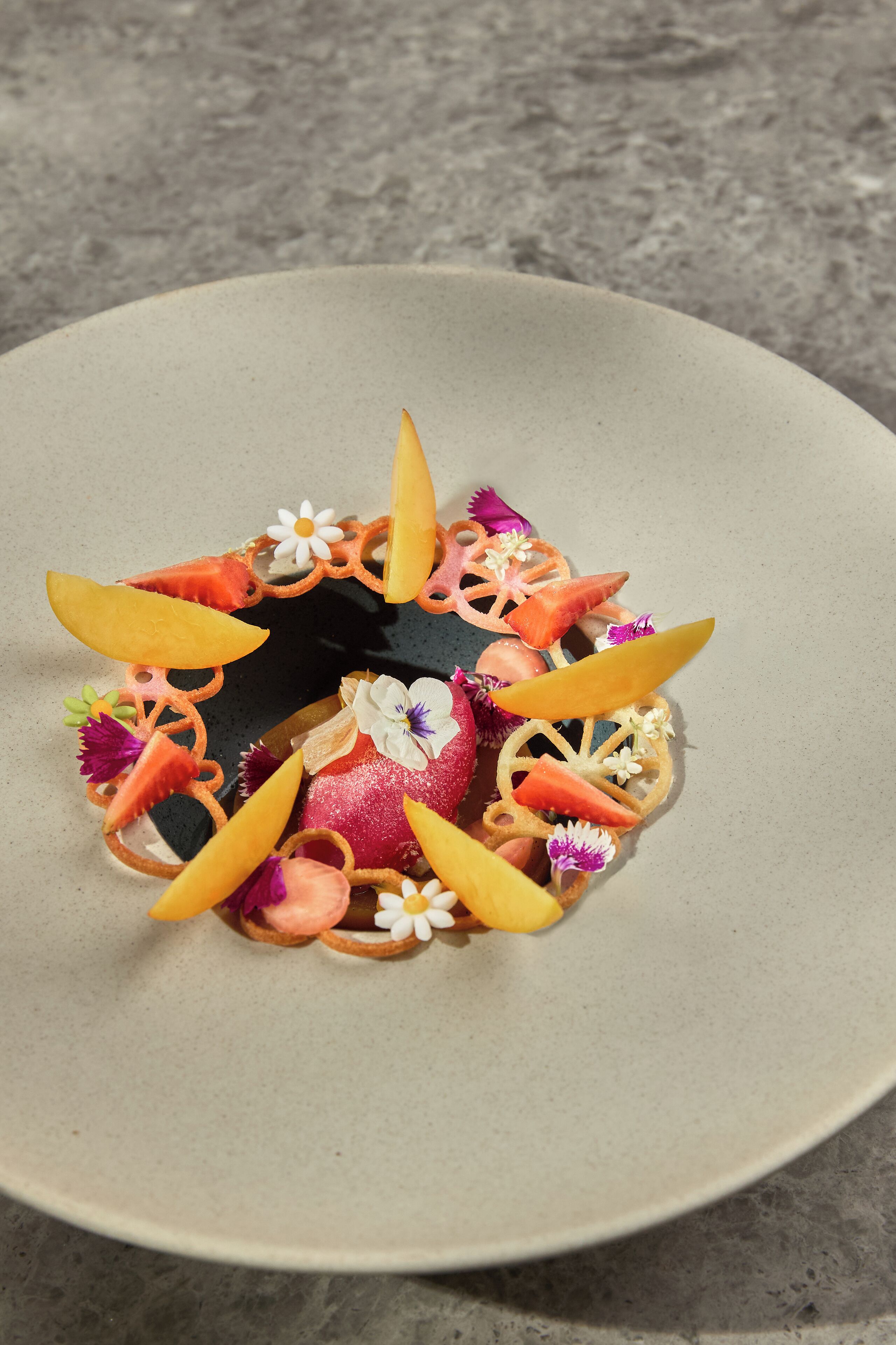 Un dessert servi dans une assiette ronde, agrémenté de divers fruits et de fleurs.