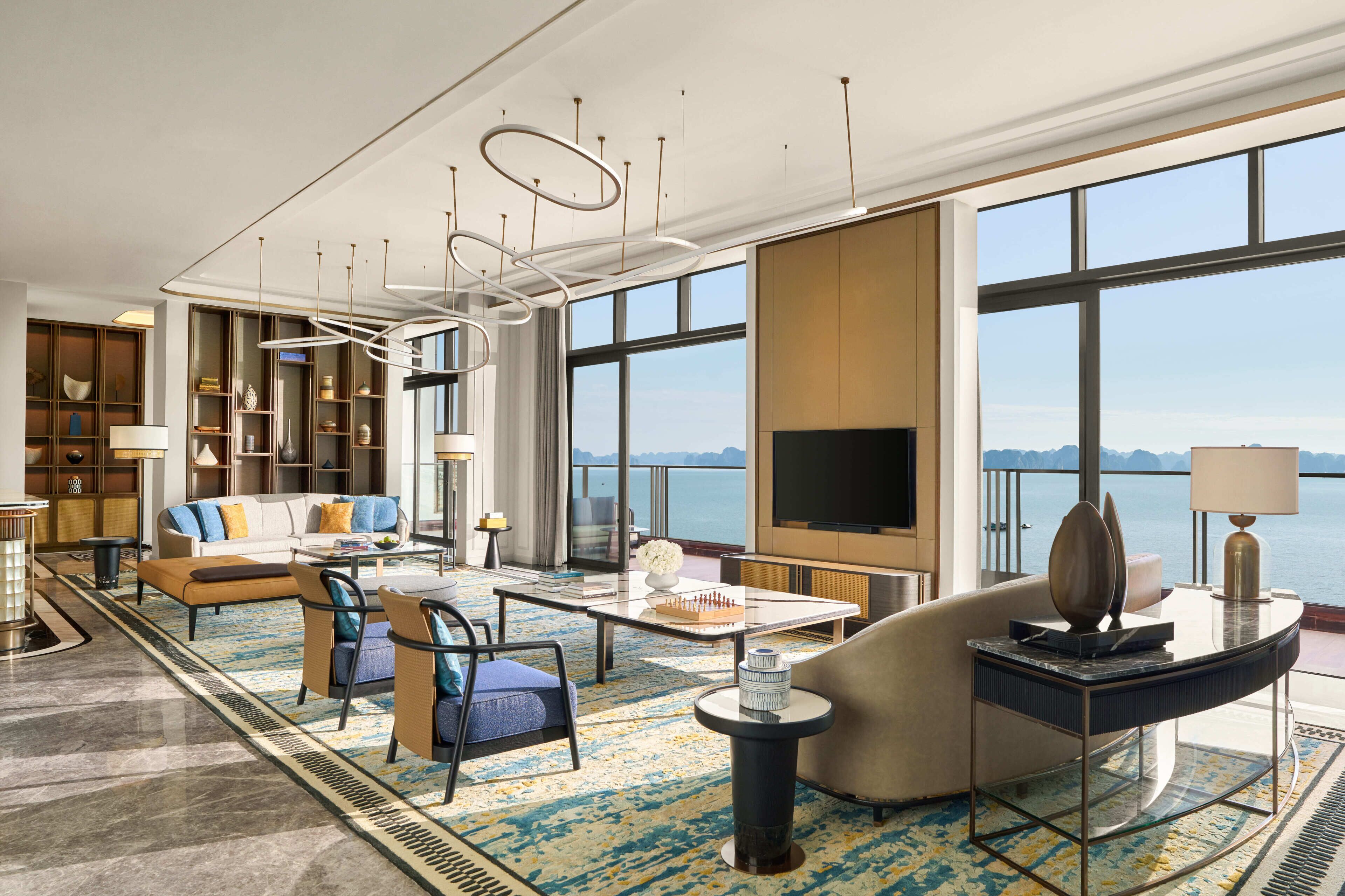 A modern hotel room with a large TV, comfortable seating, a bookshelf, and a view of the sea.