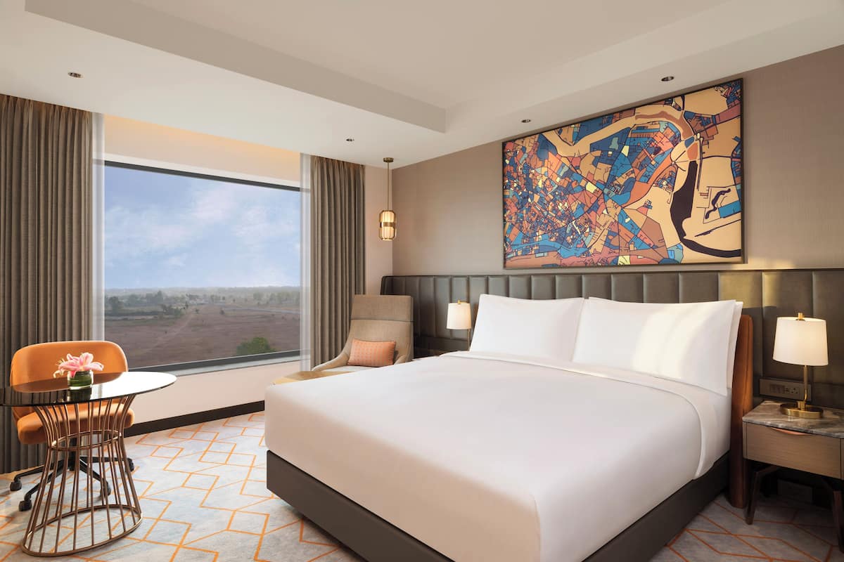 Habitación de hotel con una cama grande, un escritorio con silla, un ventanal con vistas y un cuadro de paisaje urbano en la pared.