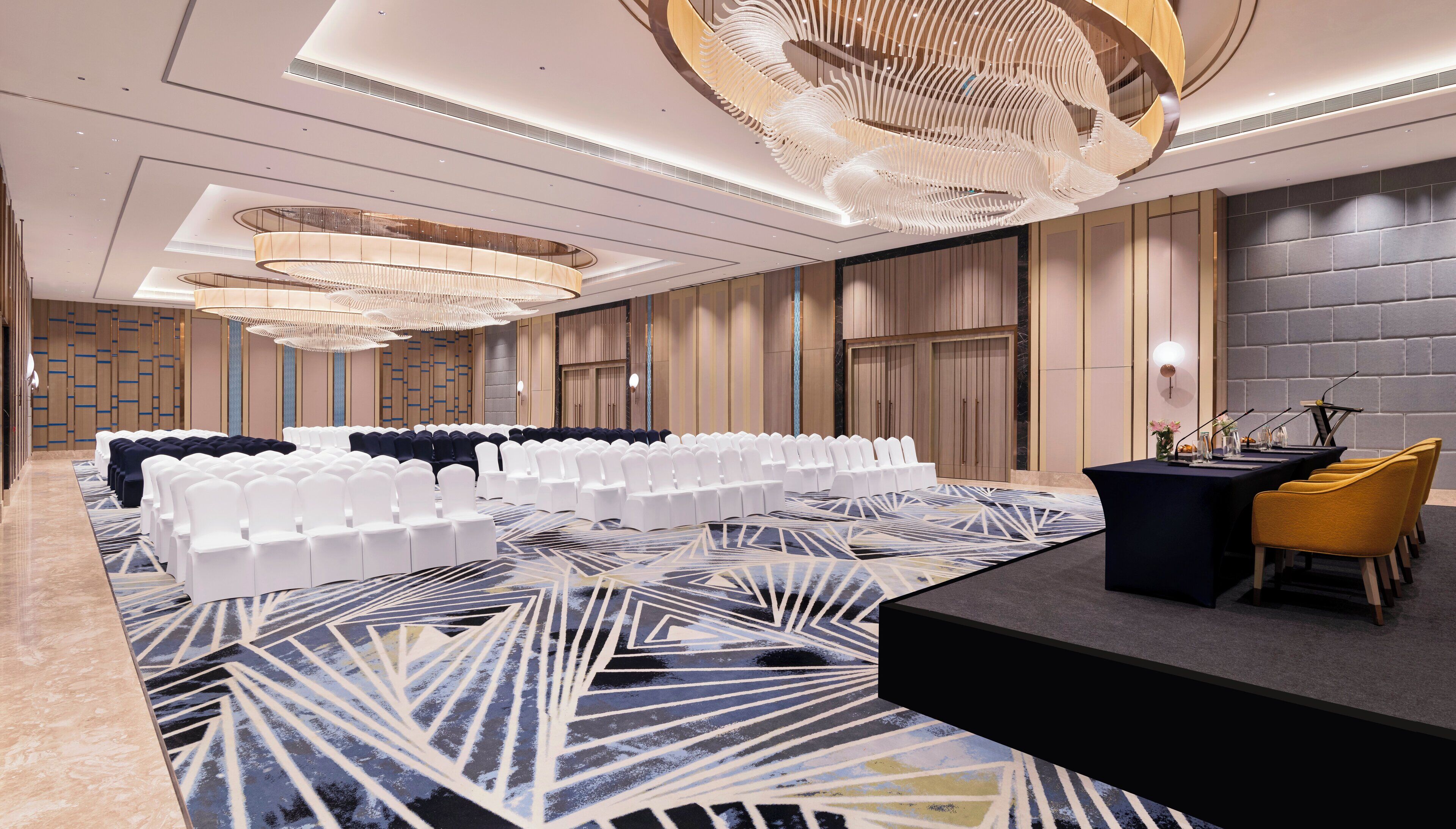 Une salle de conférence avec des rangées de chaises blanches, une scène dotée d’un fond noir et un lustre moderne.