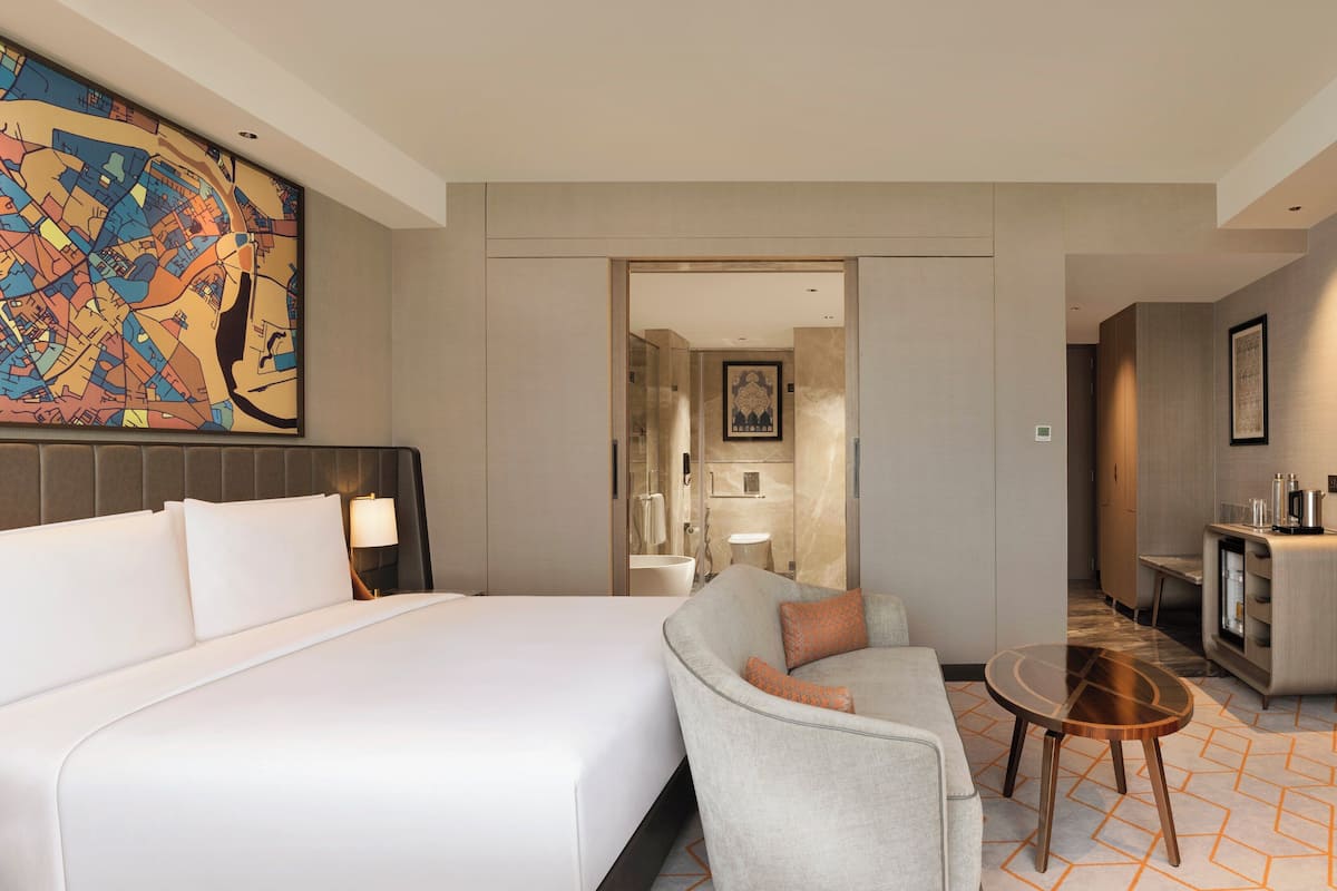 Habitación de hotel con una cama grande, un sofá, una mesita, un televisor y un baño visible a través de una puerta abierta.