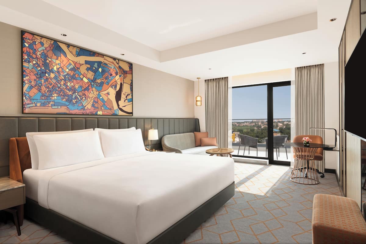 Habitación de hotel con una cama grande, un escritorio, una silla, un sofá y vistas a la ciudad a través de amplios ventanales.