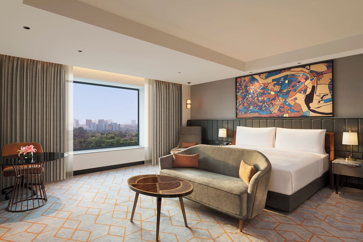 Habitación de hotel con una cama grande, un sofá, una pequeña mesa redonda, un escritorio y un amplio ventanal con vistas a la ciudad.