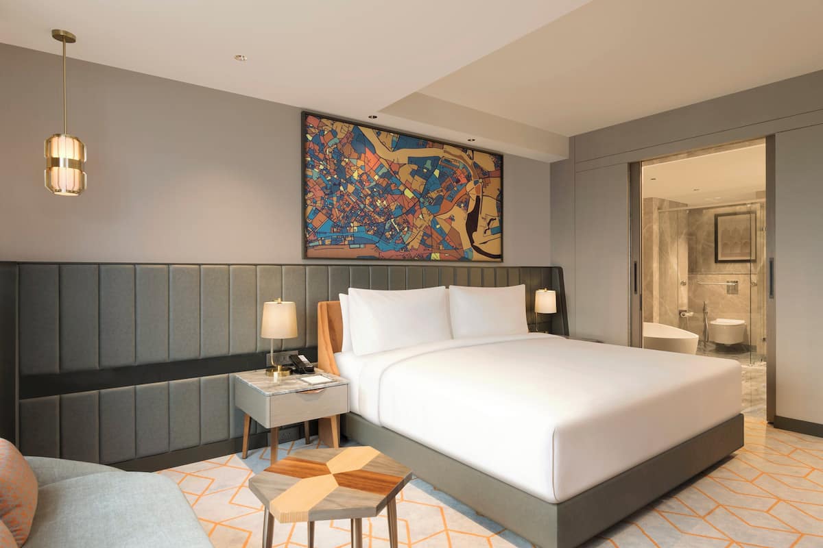 Una habitación de hotel moderna con una cama grande, una mesita de noche, un escritorio y un baño visible a través de una puerta abierta.