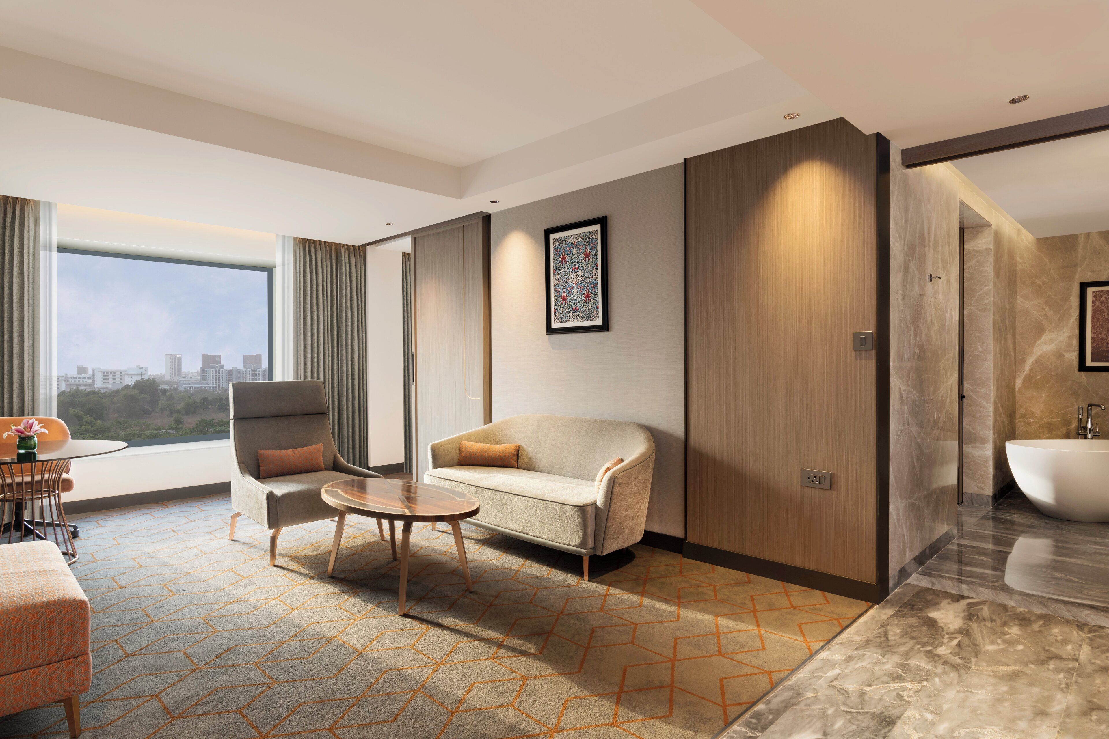 A modern hotel room with a sofa, armchair, and a small table. There is a large window with a city view, a bathroom with a bathtub, and a marble floor.