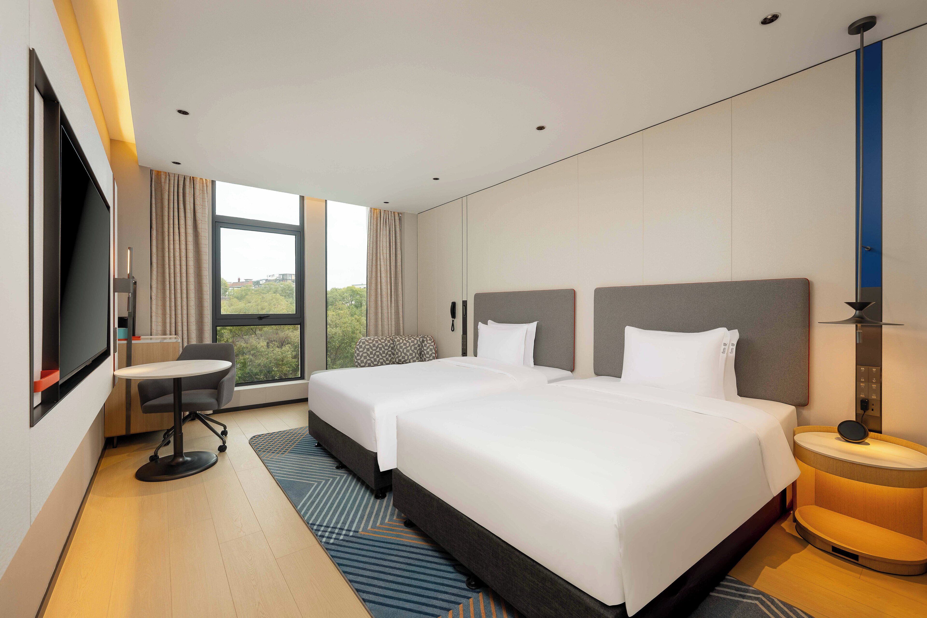 Quarto de hotel moderno com duas camas, uma escrivaninha, uma cadeira, televisão e uma janela grande com vista.